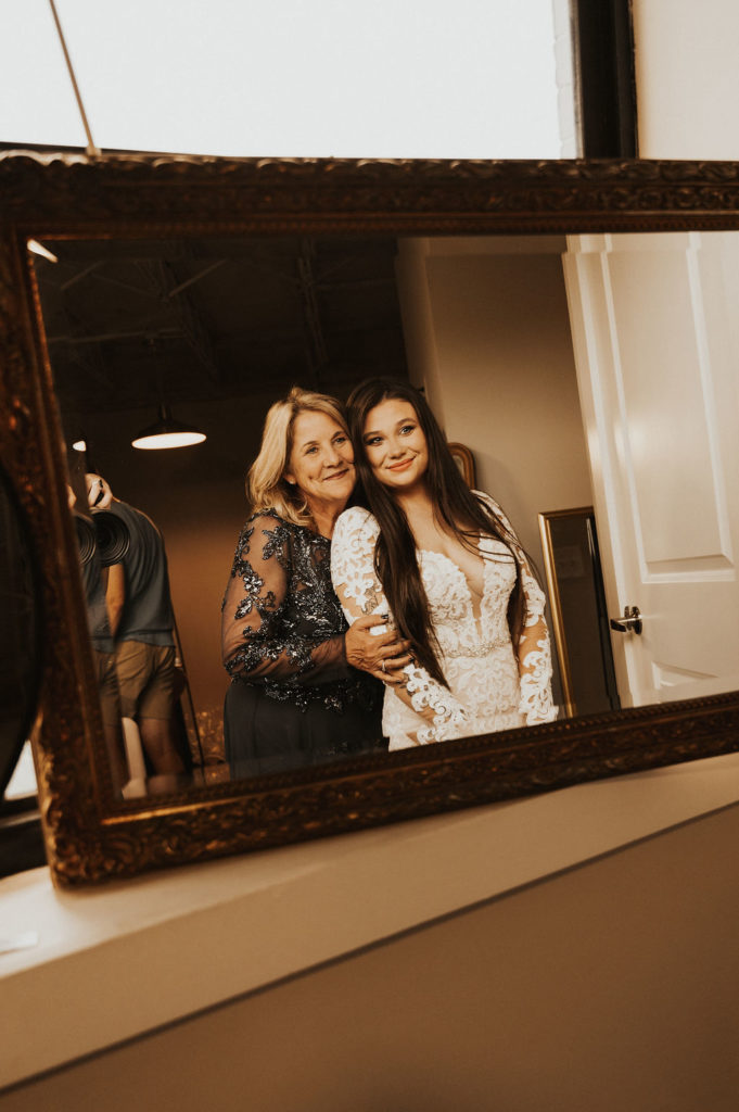 brides mother hugs her from behind as they look in the mirror