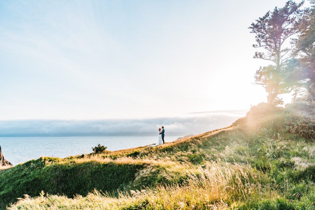 couple stands on a grassy hill overlooking the pacific ocean while the sun sets