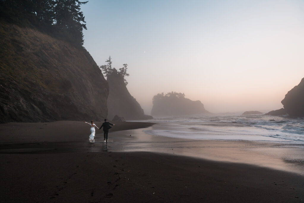 couple running barefoot across a black sand beach with their arms outstretched 