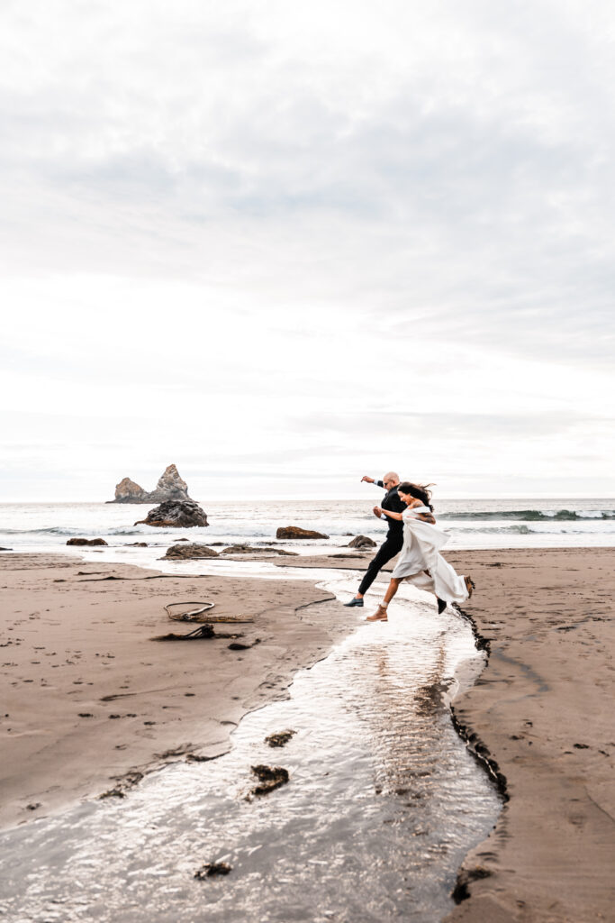 couple in wedding attire jumping over a stream