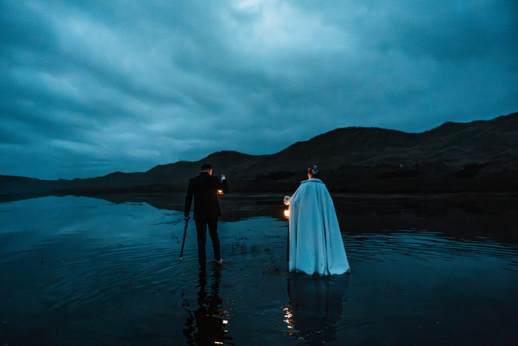 Fantasy attire couple wearing capes holding lanterns and swords walking through water