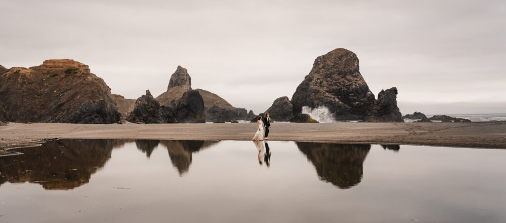 couple walking with sea stacks and crashing waves behind them.